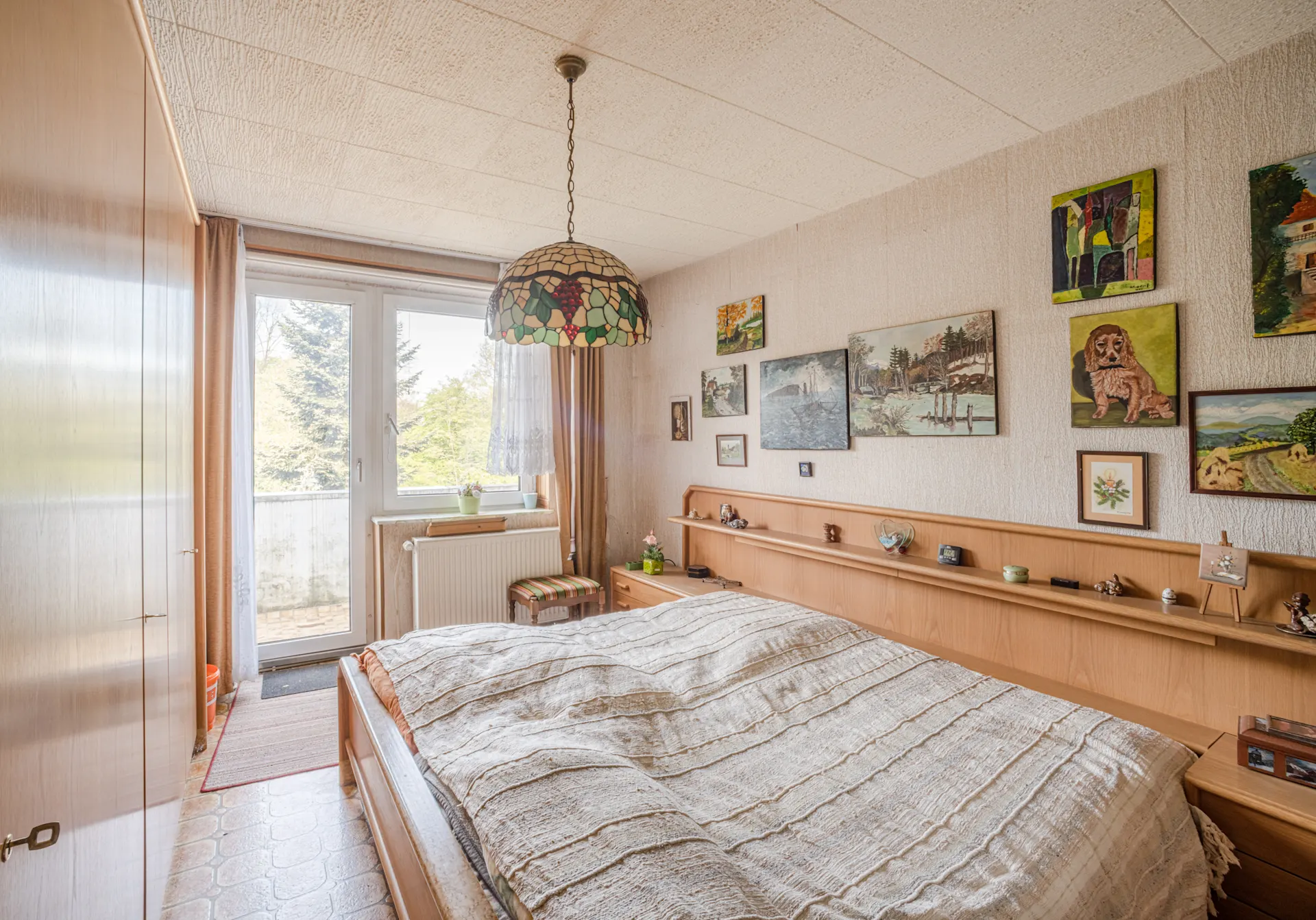 Helles Schlafzimmer mit Holzmöbeln, großem Fenster, Bett und mehreren Bildern an der Wand.