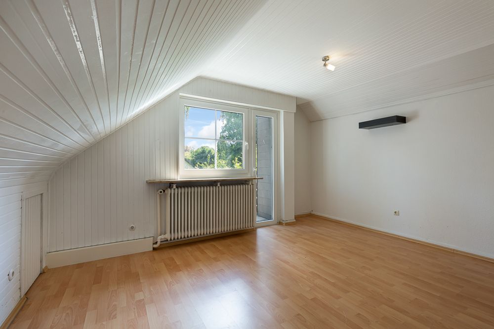 Leeres Zimmer mit Holzfußboden, Fenster, Tür zum Balkon und weißer Wandverkleidung.