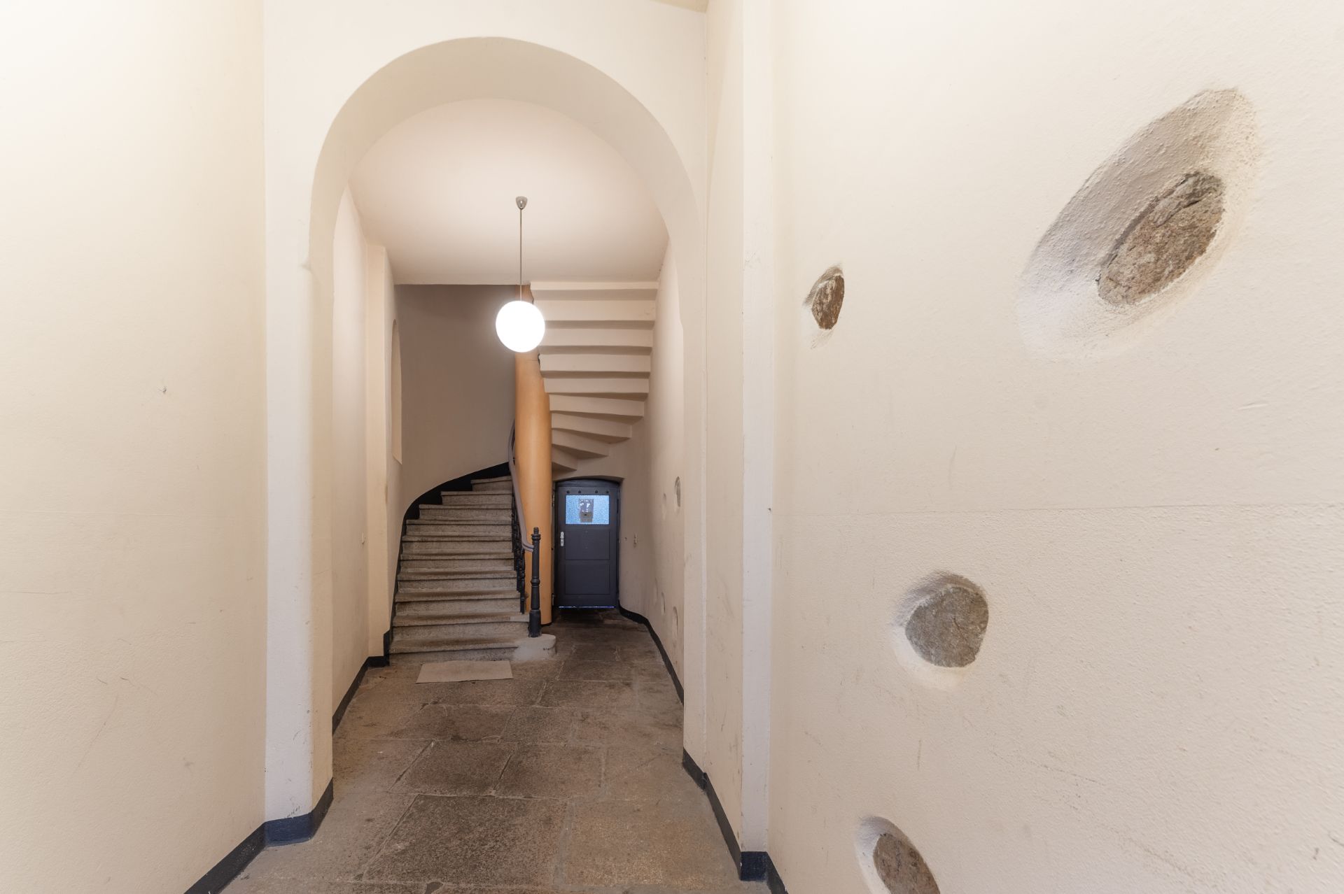 Flur mit Steinboden, runder Deckenleuchte und gewundener Treppe, Wand mit eingelassenen Steinen rechts.