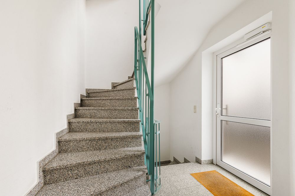 Treppenhaus mit Granitstufen, grünem Geländer und Glastür mit Matte.
