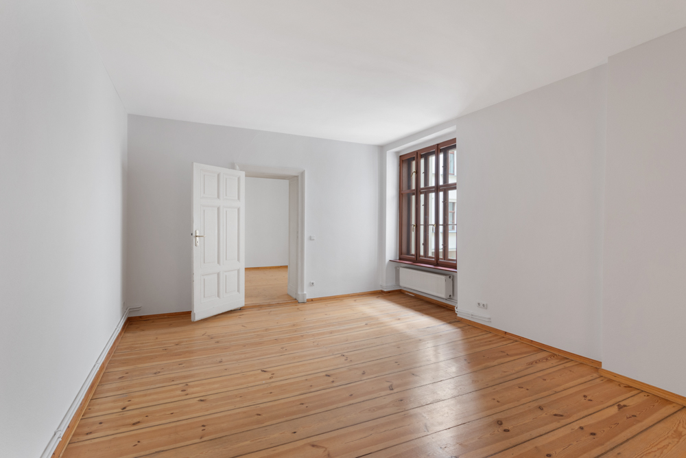 Leerer Raum mit Holzboden, weißer Wand, großem Fenster und offener Tür.