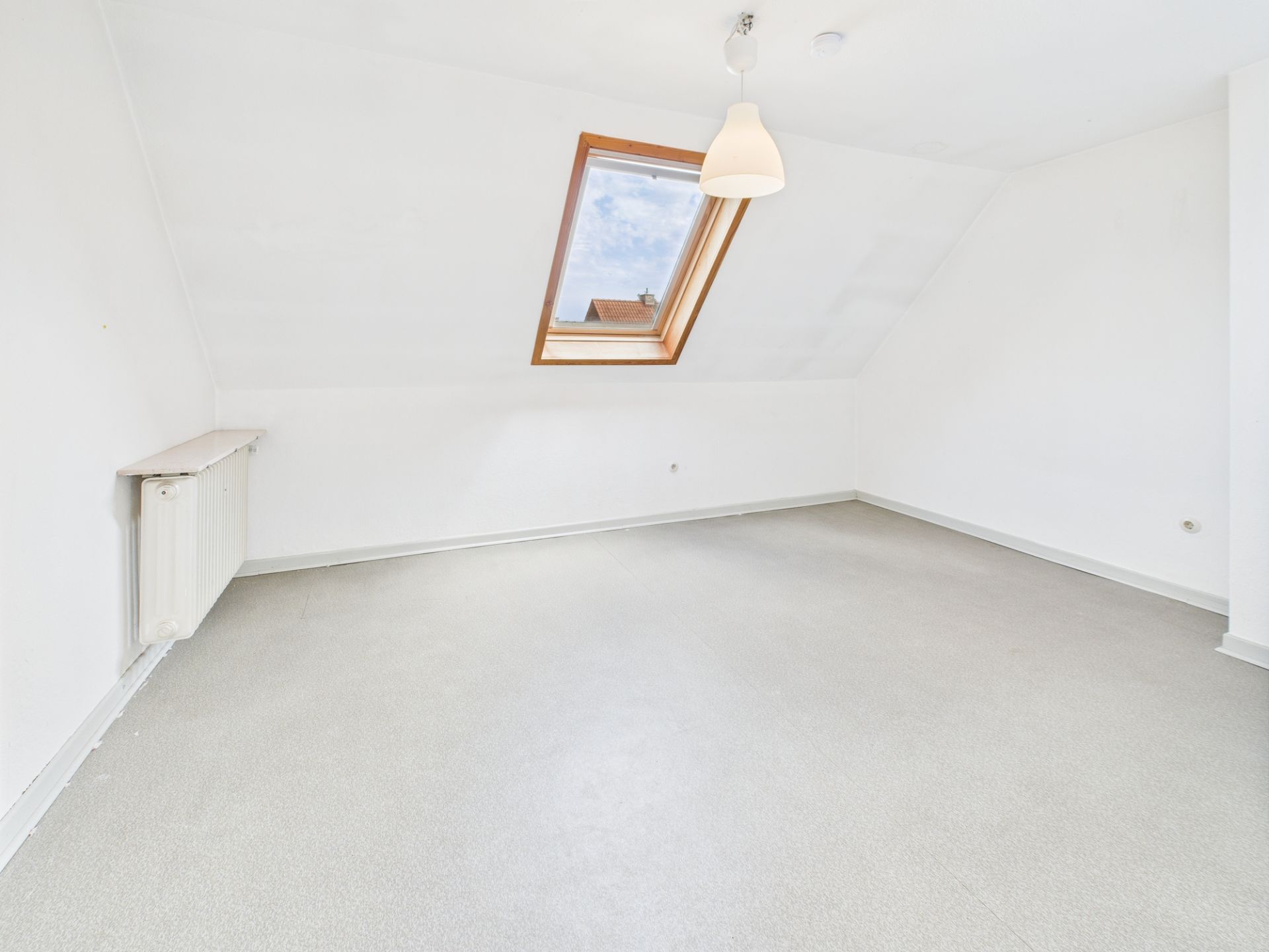 Helles, leeres Dachzimmer mit schräger Decke, Dachfenster und weißer Lampe.