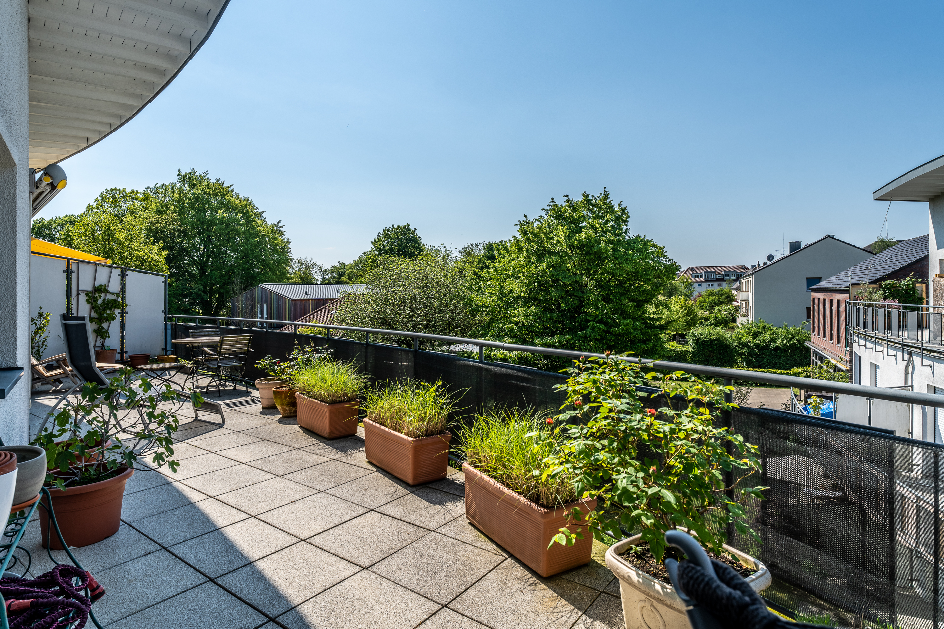 Große Terrasse mit Pflanzkübeln, Sitzmöbeln und Blick auf Bäume sowie Nachbarhäuser.