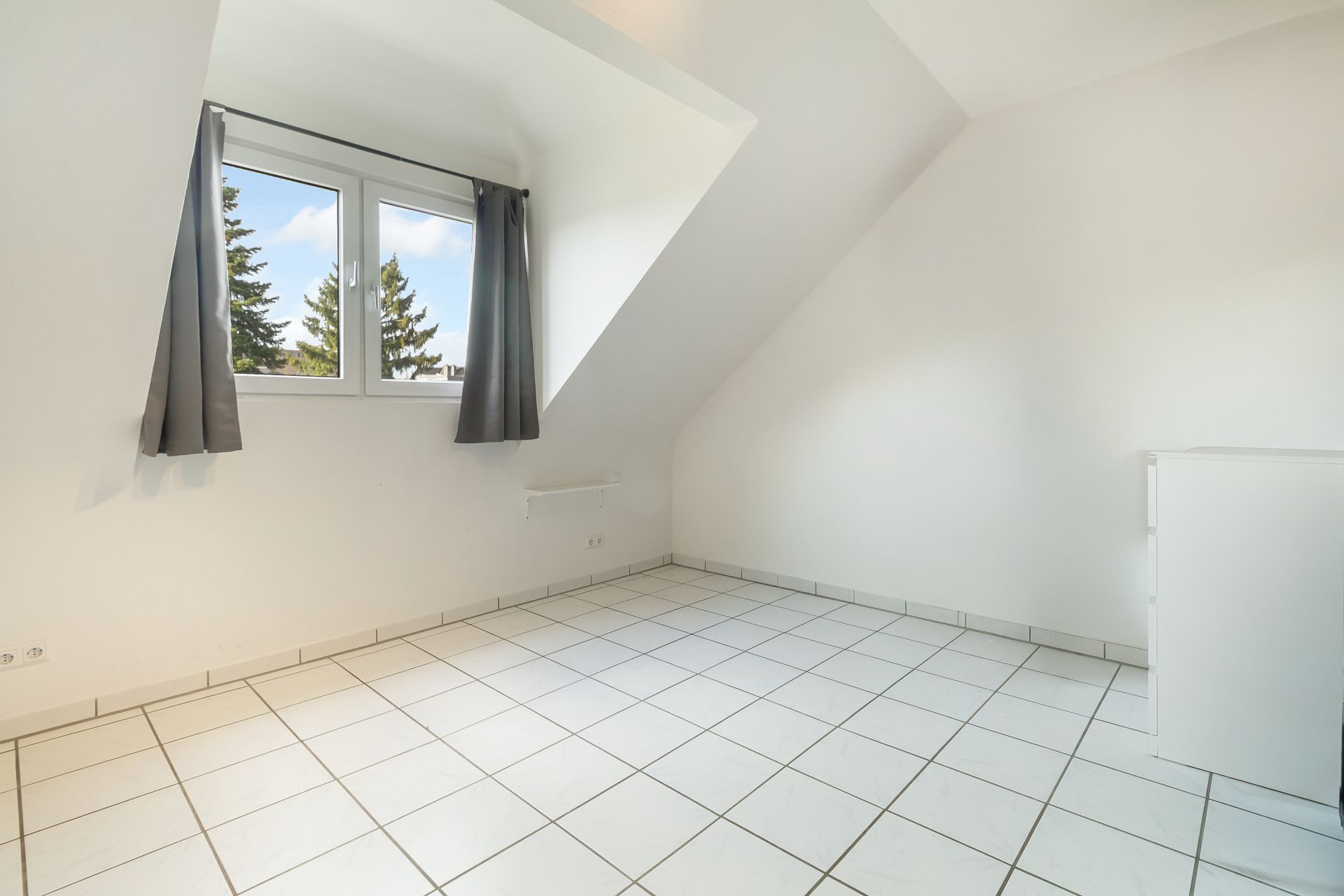 Helles Zimmer mit weißen Fliesen, Dachschräge und Fenster mit grauen Vorhängen.