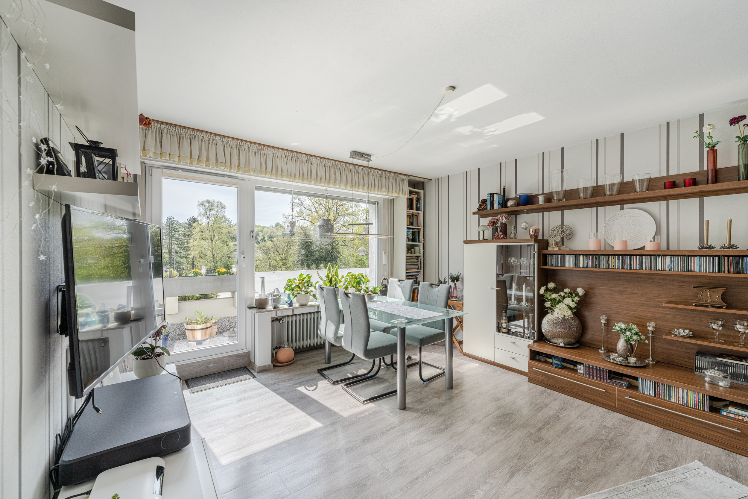 Helles Wohnzimmer mit Glastisch, Stühlen, Regalwand, TV und Balkon mit Pflanzen.