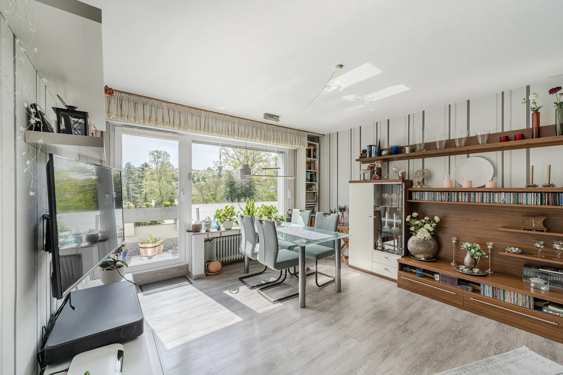 Helles Wohnzimmer mit Glastisch, Stühlen, Regalwand, TV und Balkon mit Pflanzen.