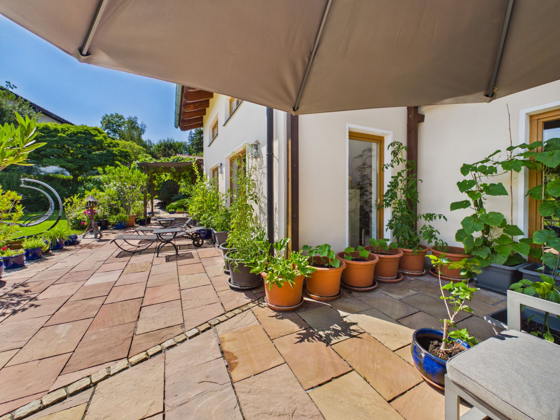 Sonnige Terrasse mit vielen Blumentöpfen, Gartenmöbeln und Blick ins Grüne.