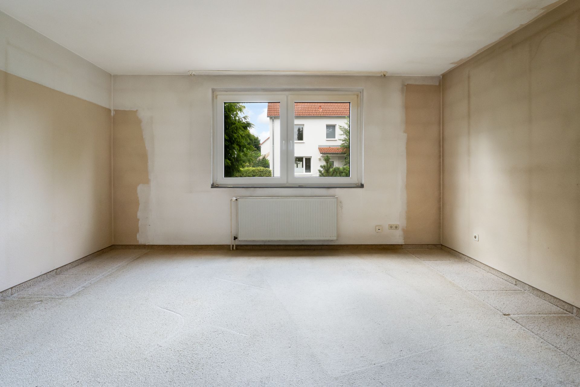 Leerer Raum mit Teppichboden, großem Fenster und Heizkörper an der Wand.