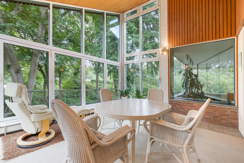 Wintergarten mit runden Tisch, Korbstühlen und Sessel, große Fenster mit Blick auf Bäume.