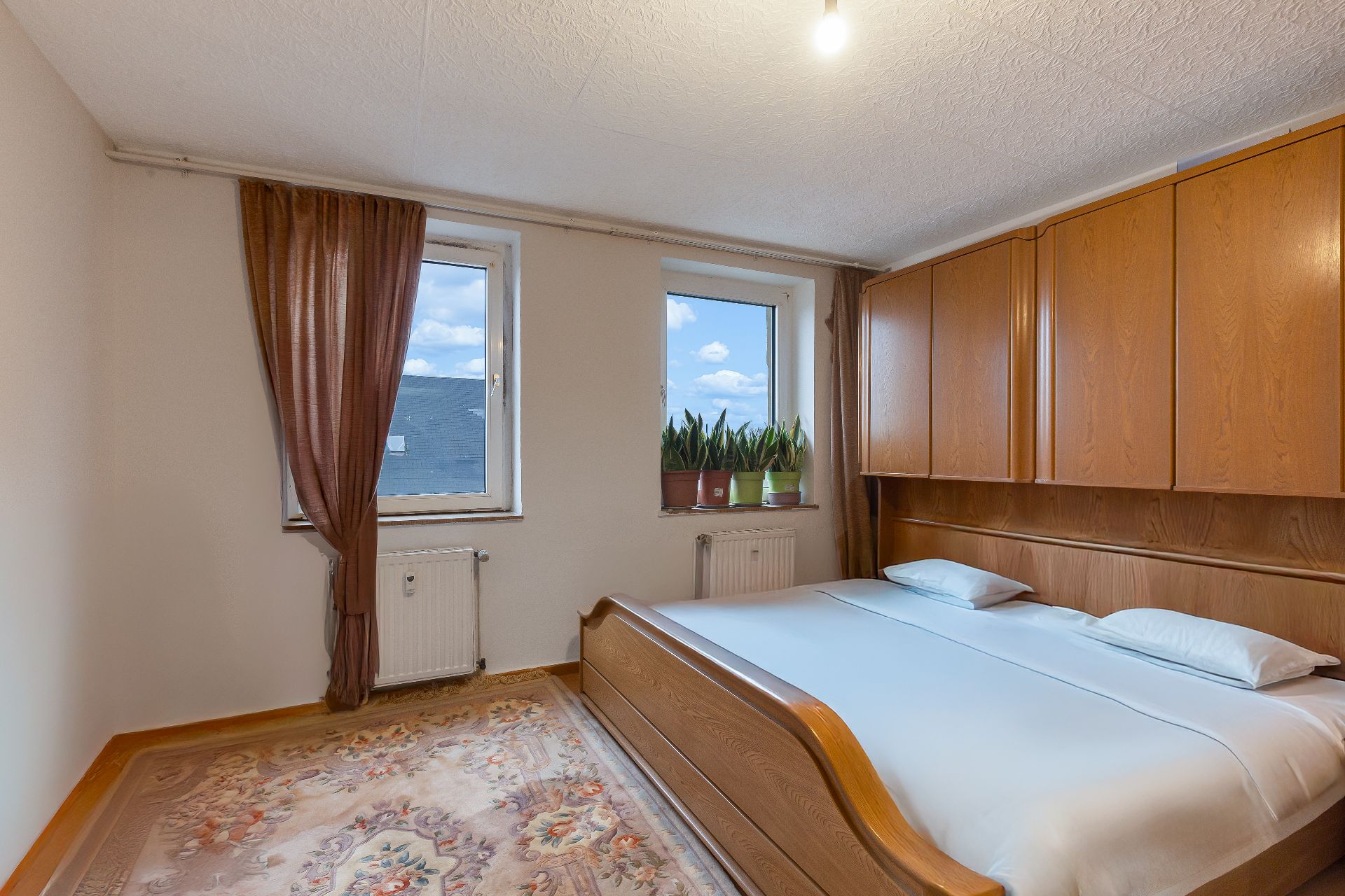Schlafzimmer mit Doppelbett, Holzmöbeln, Teppich und zwei Fenstern mit Vorhängen und Pflanzen.