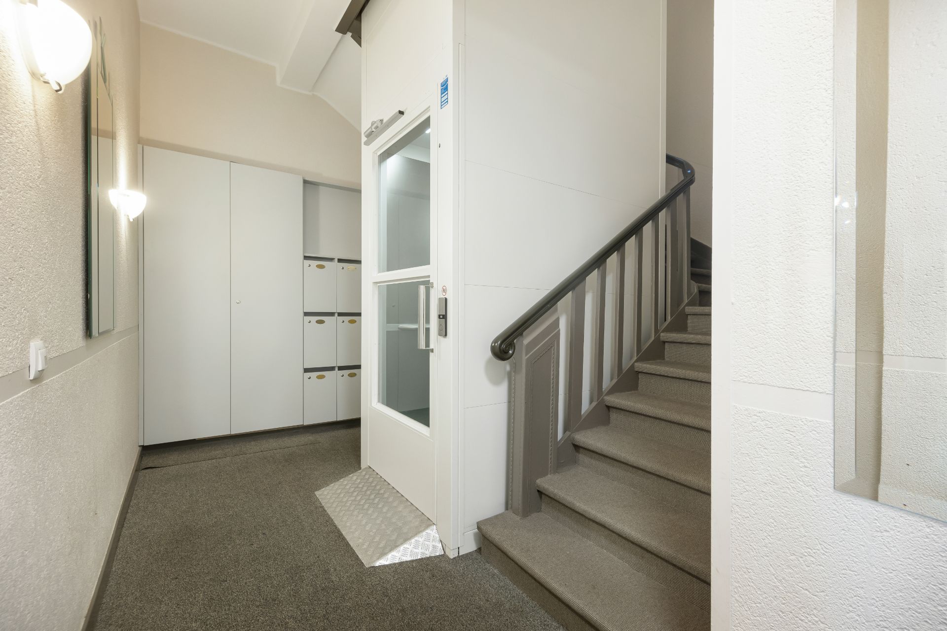 Heller Flur mit Aufzug, Treppe und weißen Schränken in einem Wohnhaus.