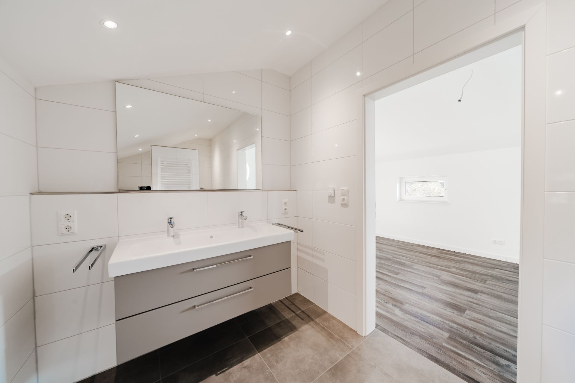 Modernes Bad mit zwei Waschbecken, großem Spiegel und Blick auf ein angrenzendes Zimmer mit Holzfußboden.