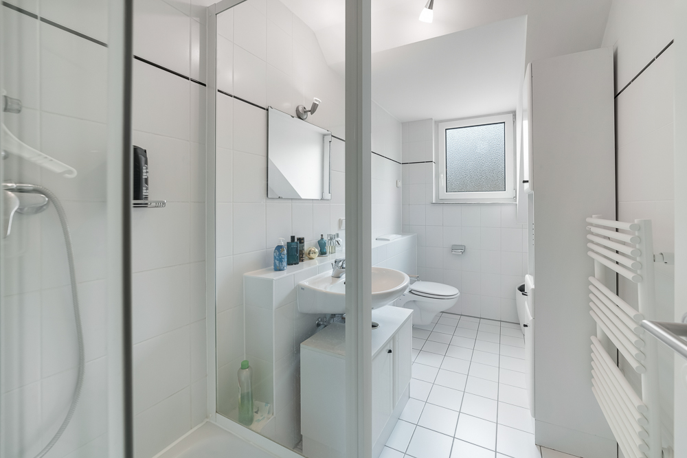 Modernes, weiß gefliestes Badezimmer mit Dusche, Waschbecken, Spiegel, WC und Fenster.