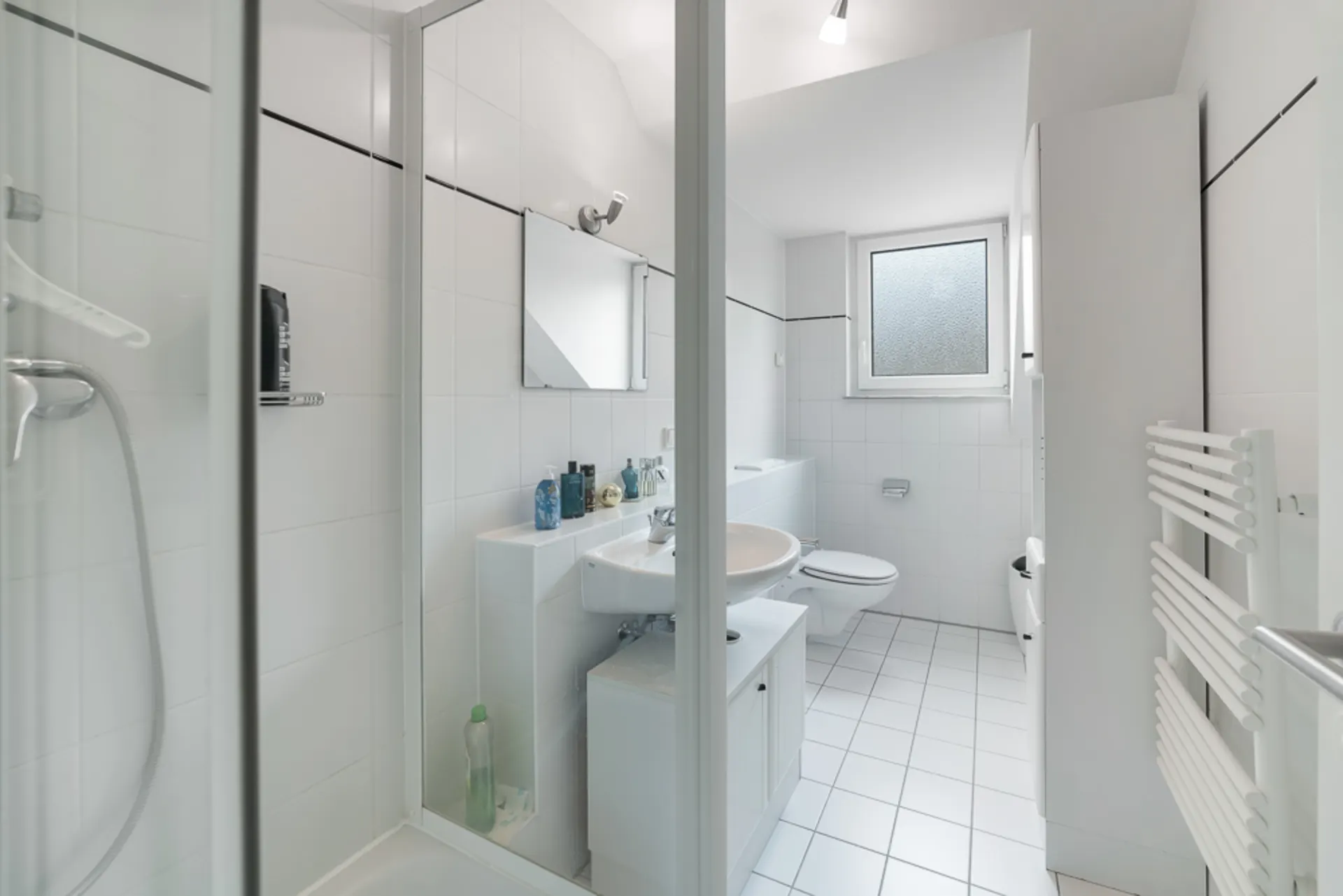 Modernes, weiß gefliestes Badezimmer mit Dusche, Waschbecken, Spiegel, WC und Fenster.