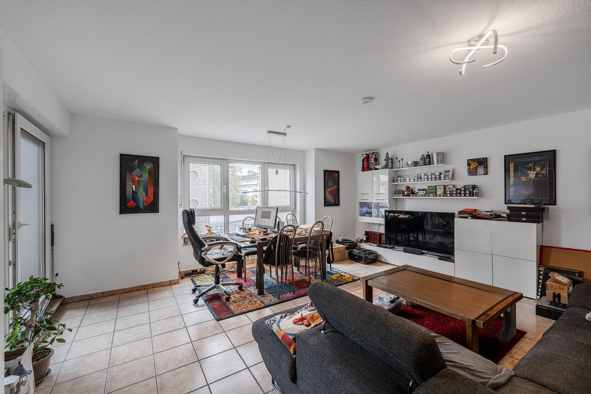 Helles Wohnzimmer mit Sofa, Schreibtisch, Regalwand, Fernseher und großen Fenstern.