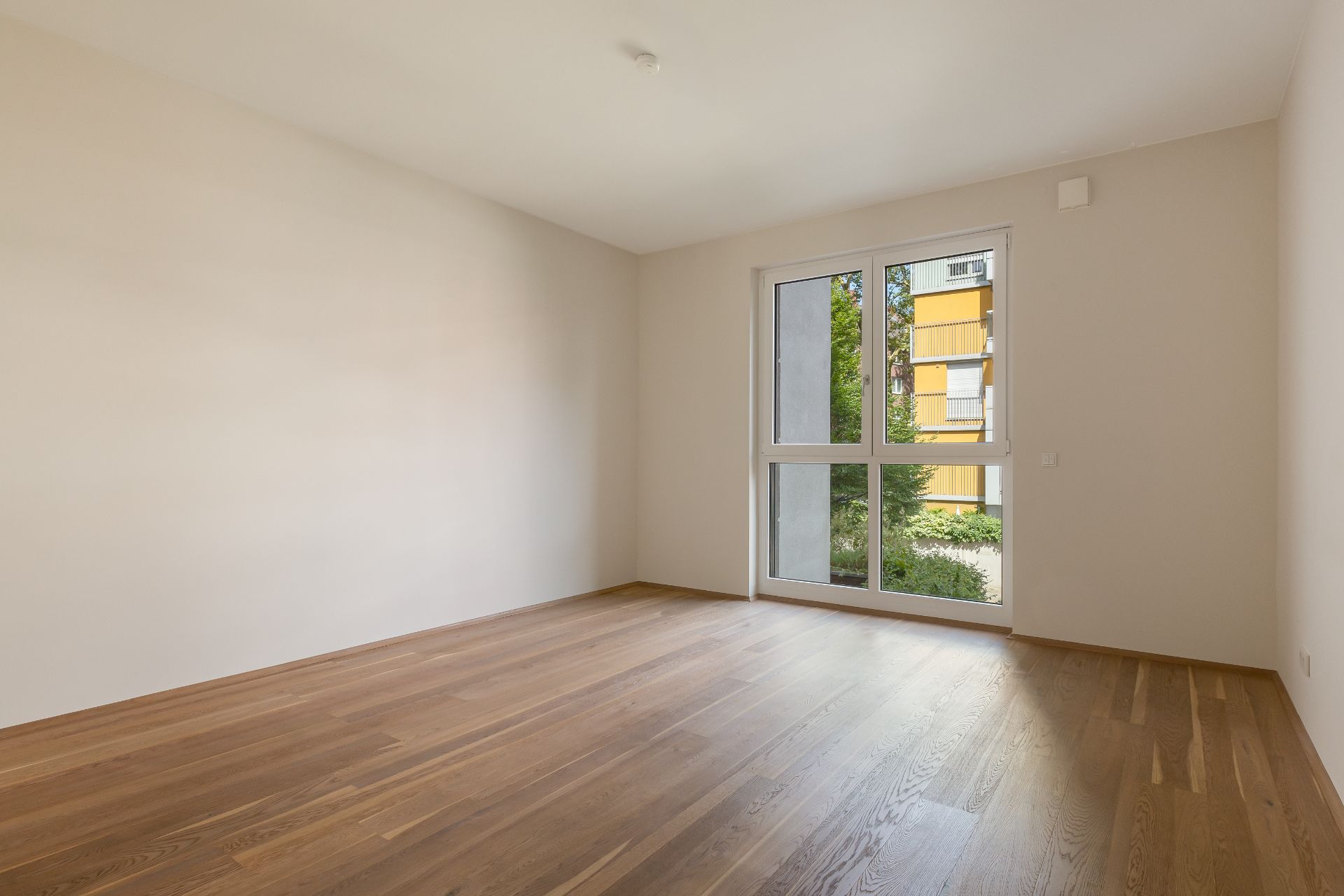 Leerer Raum mit Holzfußboden und großem Fenster, Blick auf Garten und Wohngebäude.