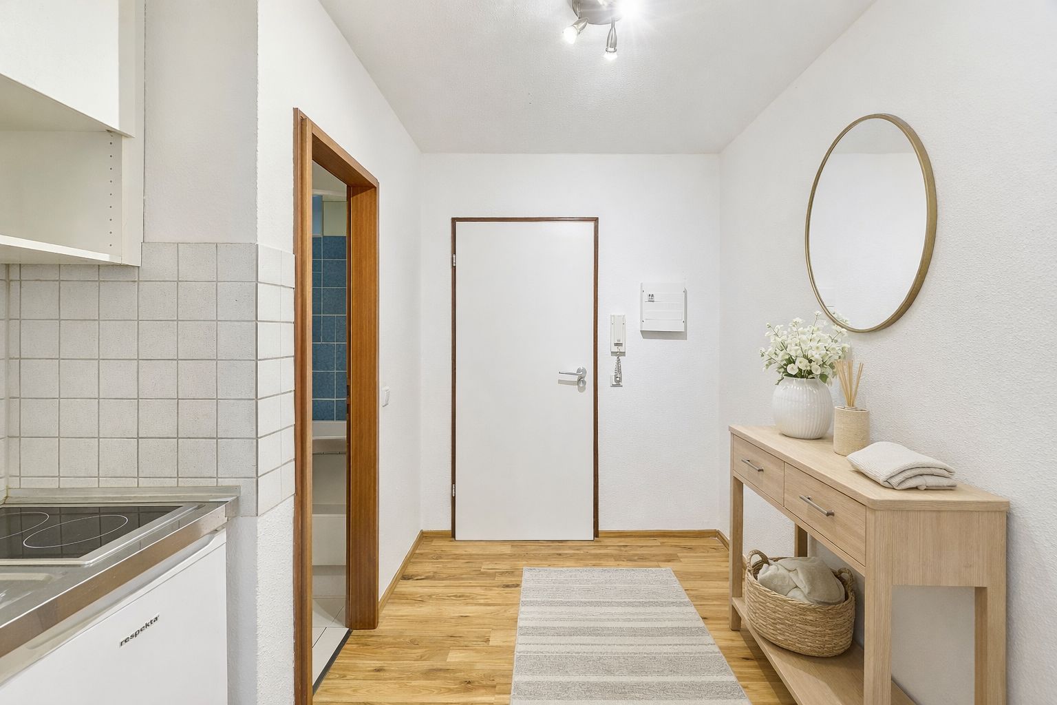 Heller Flur mit Holzboden, Kommode, rundem Spiegel und Tür, daneben Küchenbereich sichtbar.