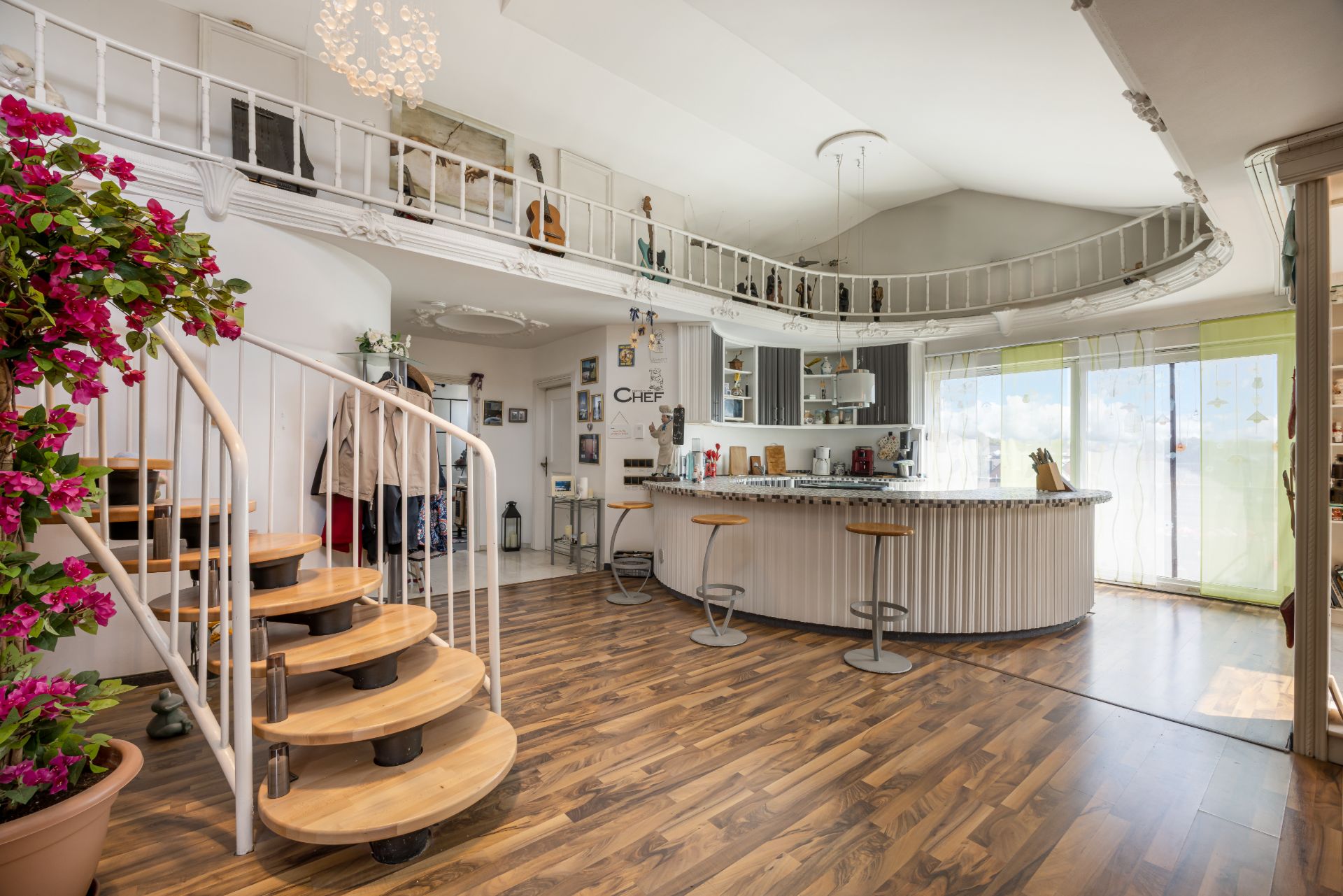 Helles Wohnzimmer mit offener Küche, Holztreppe, Bartresen und Balkon mit großen Fenstern.