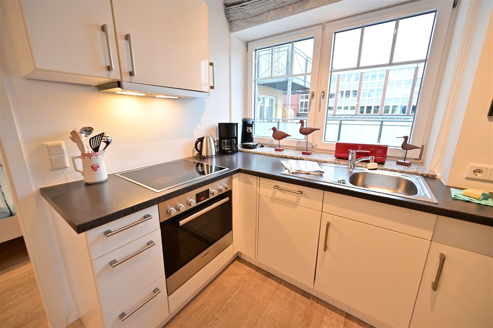Moderne, helle Einbauküche mit Herd, Spüle und Utensilien vor einem großen Fenster.