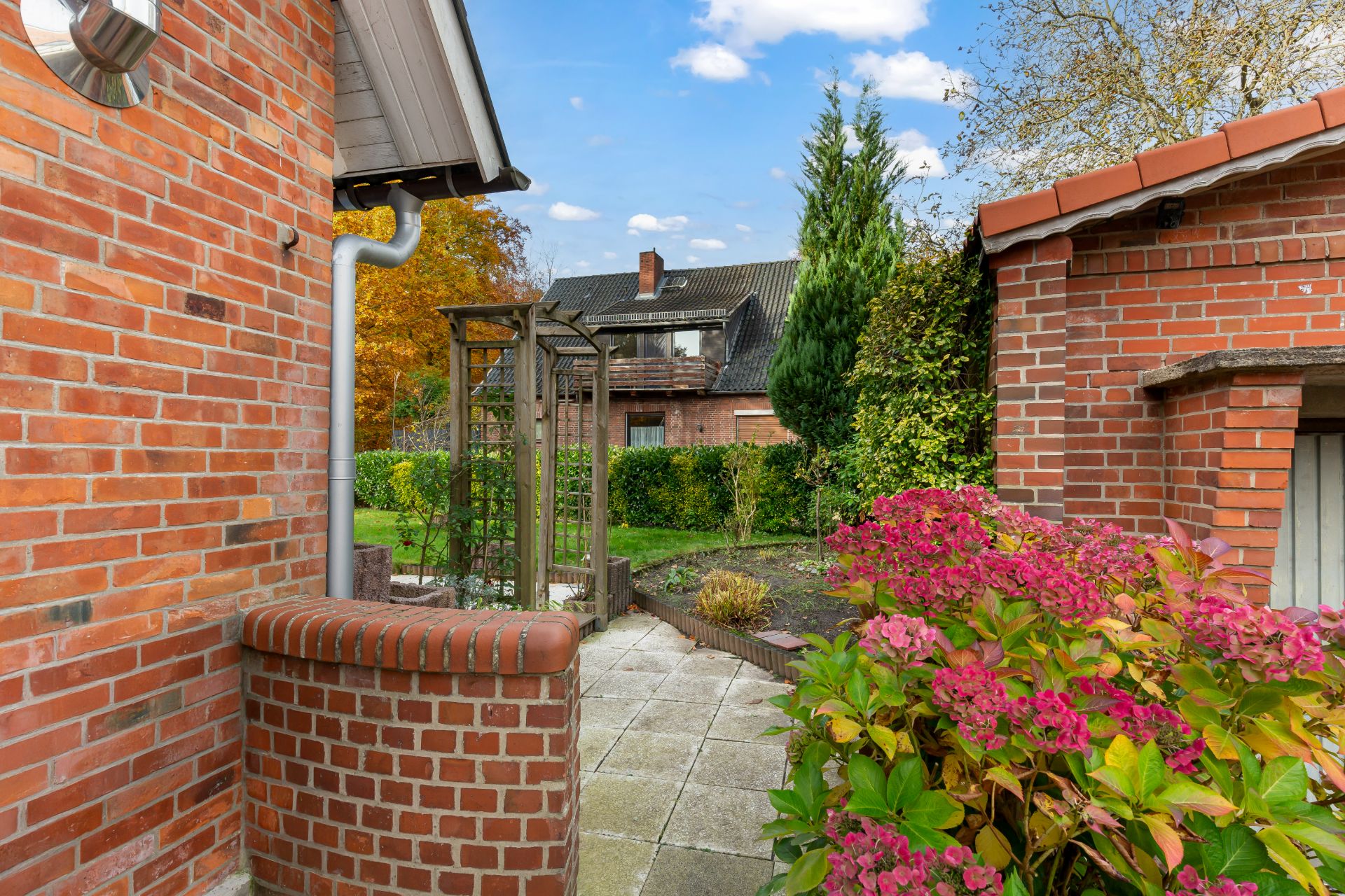 Gepflasterter Gartenbereich mit Ziegelmauern, Blumen und grünen Büschen neben einem Wohnhaus.