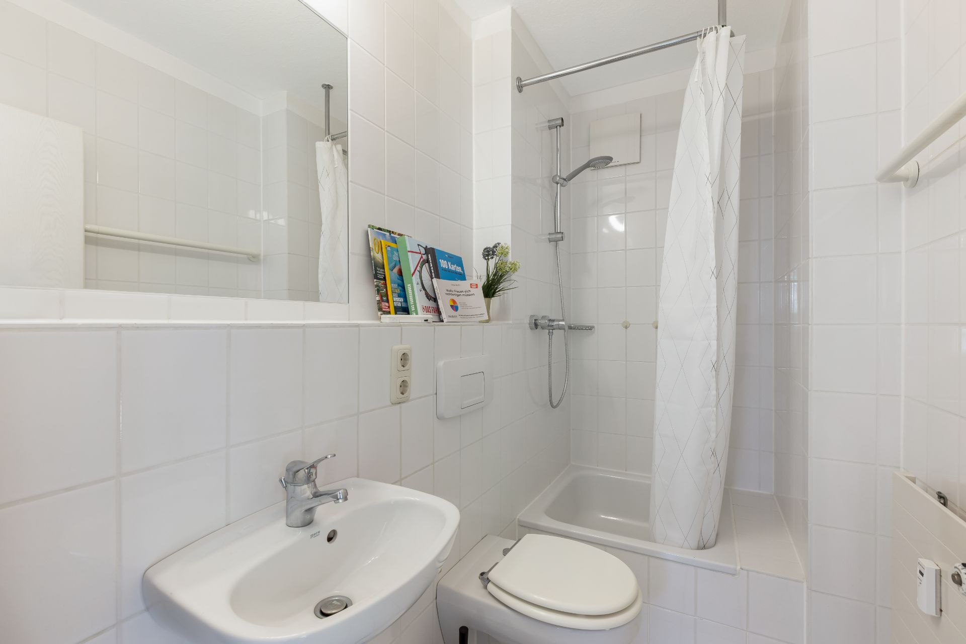 Kleines Badezimmer mit Dusche, weißem Waschbecken und Toilette, alles gefliest.