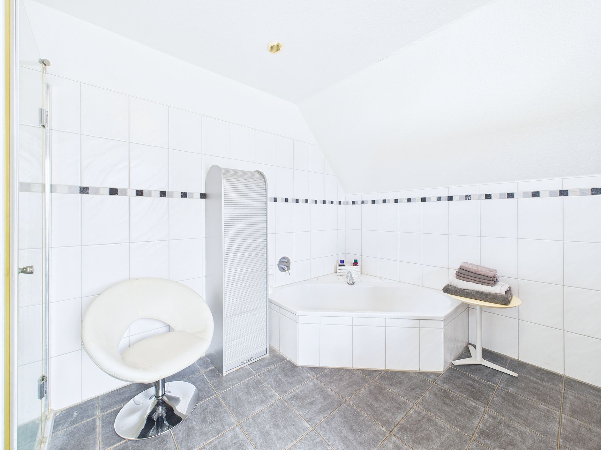 Modernes weißes Badezimmer mit Eckbadewanne, Stuhl und kleinem Tisch mit Handtüchern.