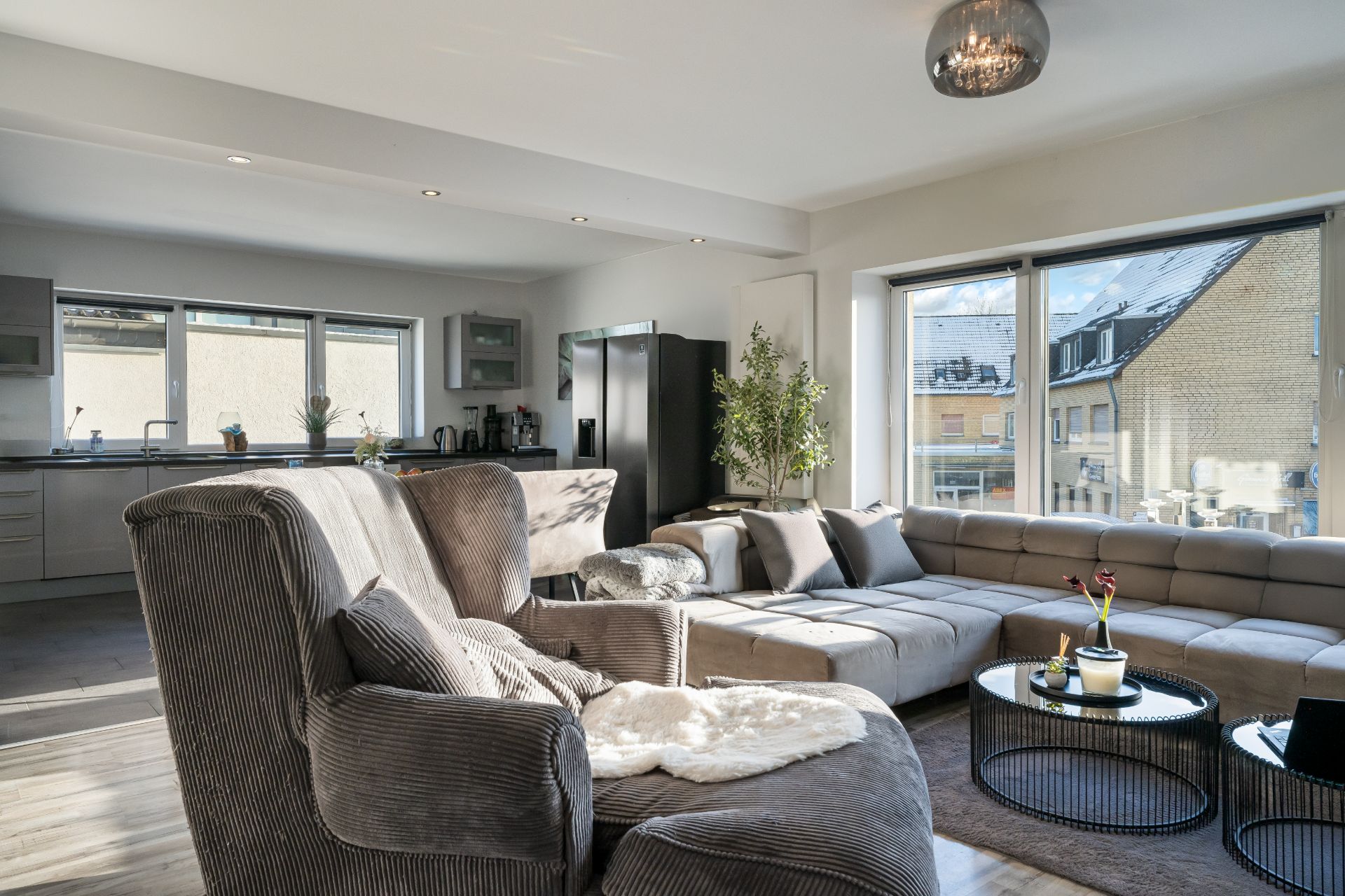 Modernes Wohnzimmer mit großer Eckcouch, Sesseln und offener Küche, viel Tageslicht durch große Fenster.