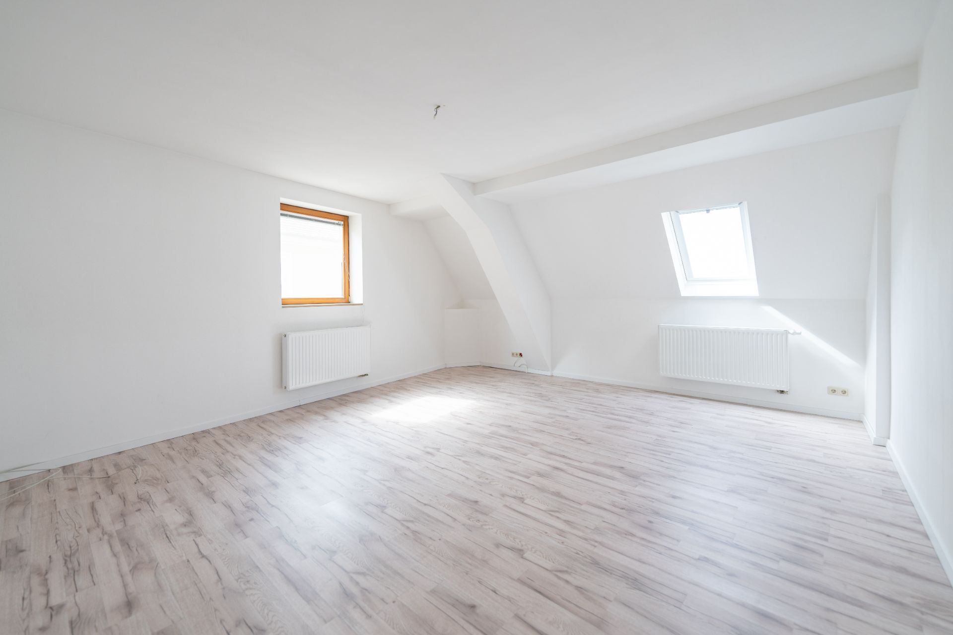 Heller, leerer Raum mit zwei Fenstern, weißen Wänden und hellgrauem Laminatboden.
