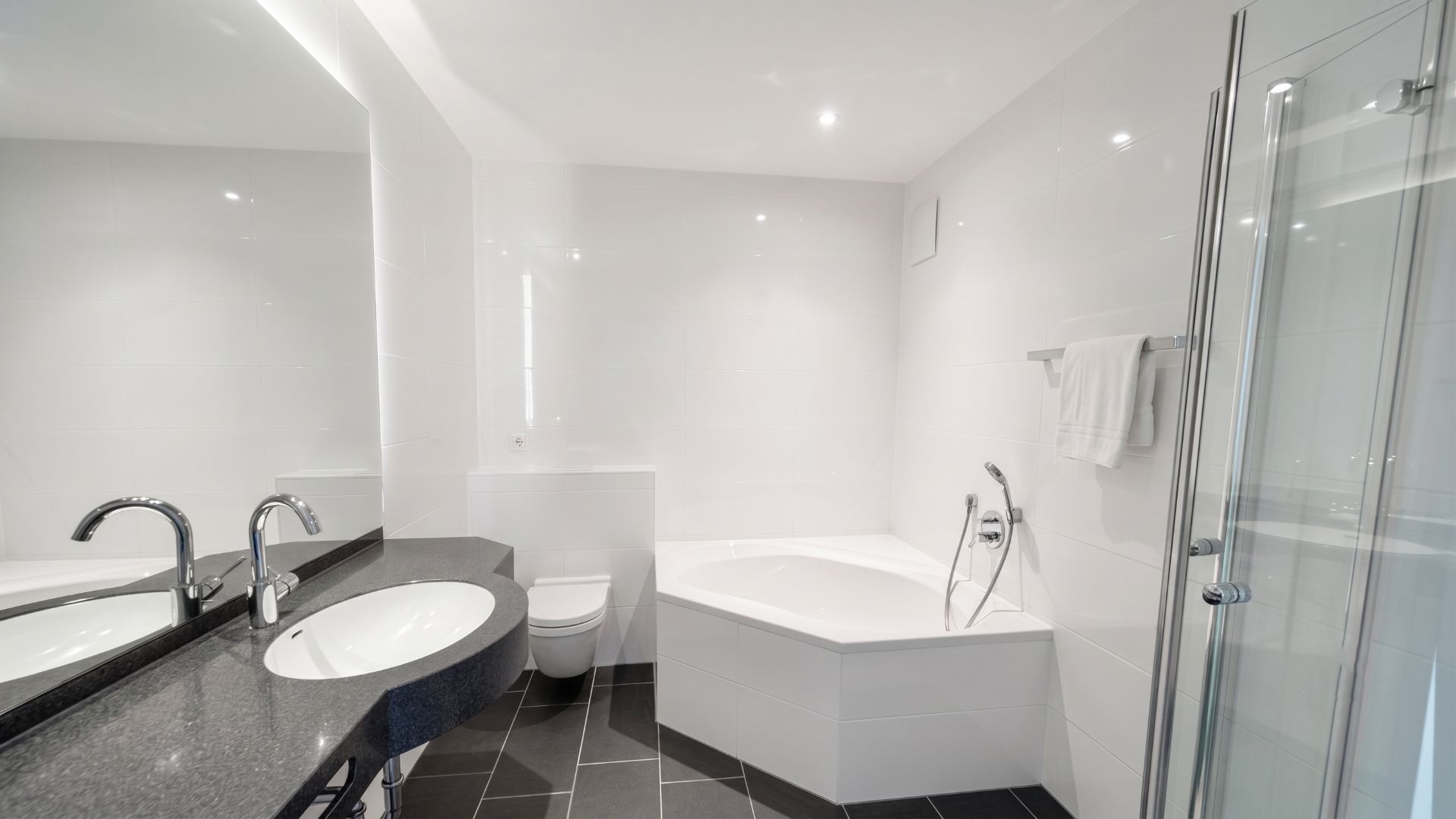 Modernes Badezimmer mit Eckbadewanne, Waschbecken, Spiegel, WC und Dusche auf dunklem Fliesenboden.