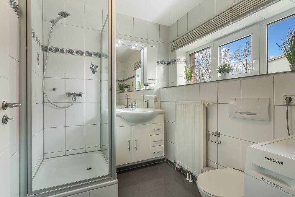 Modernes Badezimmer mit Dusche, Waschbecken, WC, Waschmaschine und Fenstern mit Pflanzen.