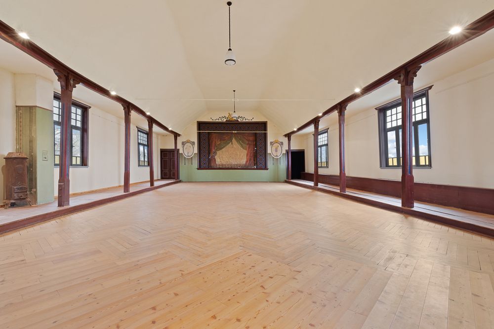 Großer Saal mit Holzboden, Bühne am Ende, hohen Fenstern und Holzbalken an den Seiten.