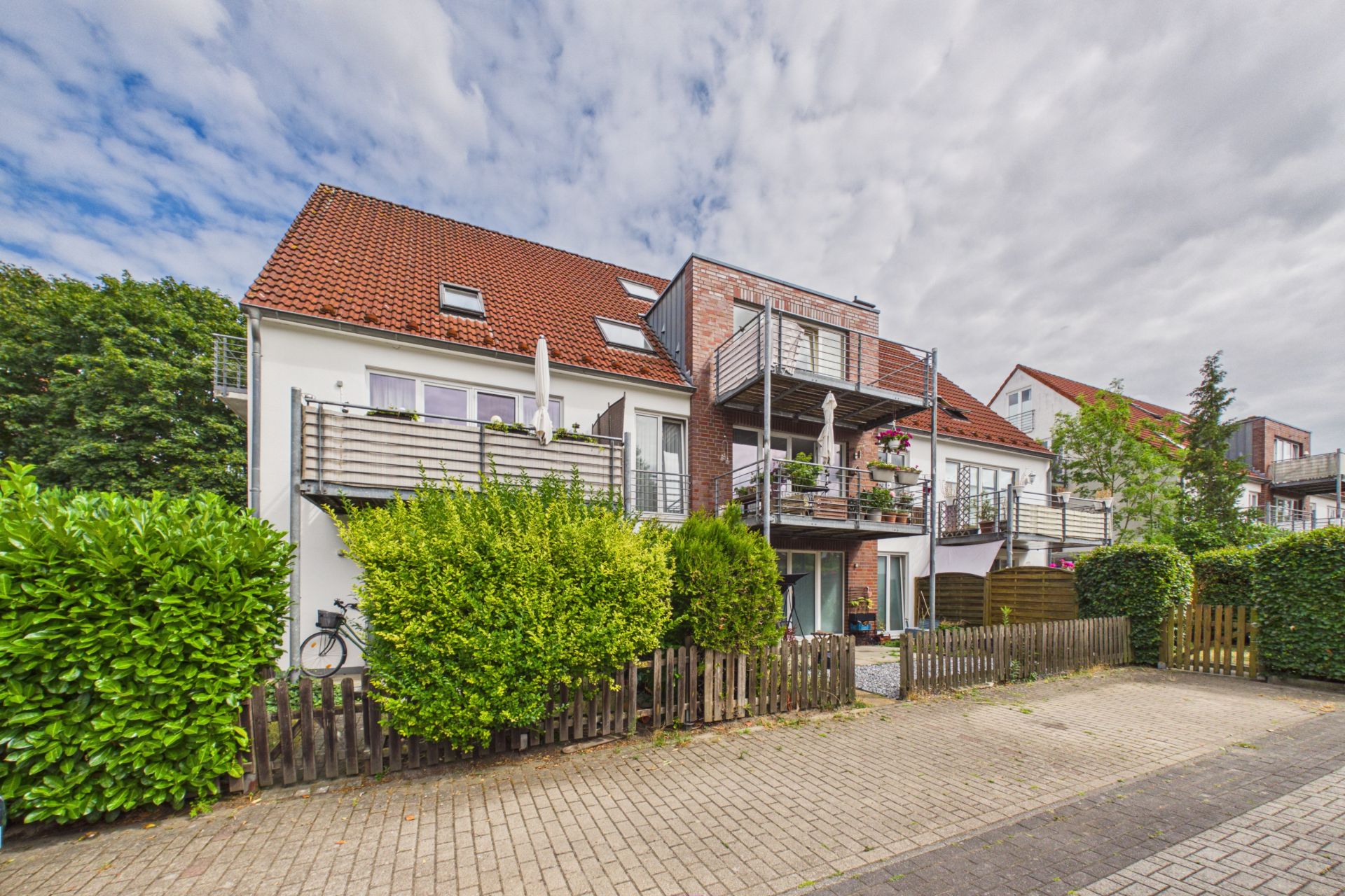 Modernes Wohnhaus mit Balkonen, roten Dachziegeln und Vorgarten, aufgenommen von der Straße.