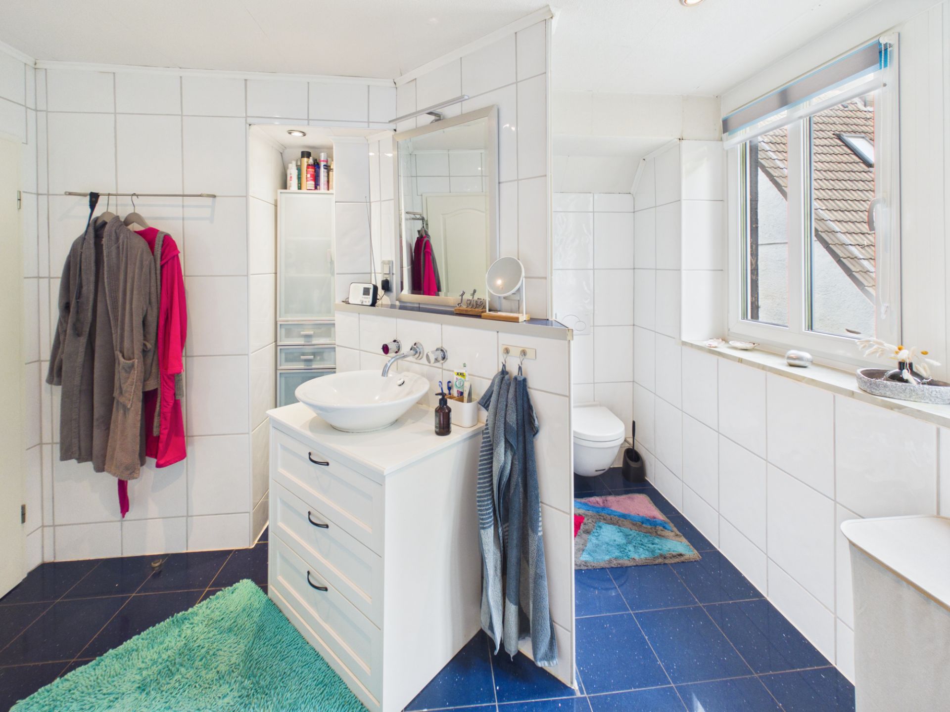 Modernes Badezimmer mit Fliesen, Waschbecken, Spiegel, Kleiderschrank und zwei bunten Teppichen.