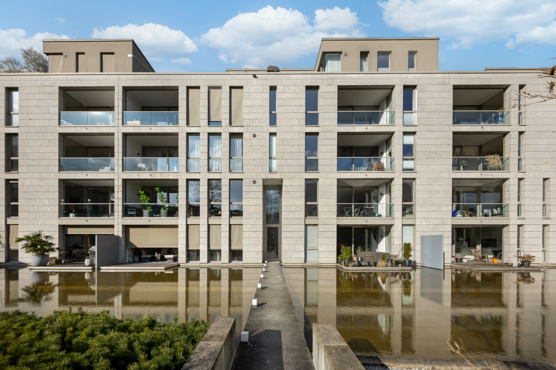 Modernes Wohnhaus mit Balkonen und Glasgeländern, davor ein flacher Wasserbereich und Gehweg.