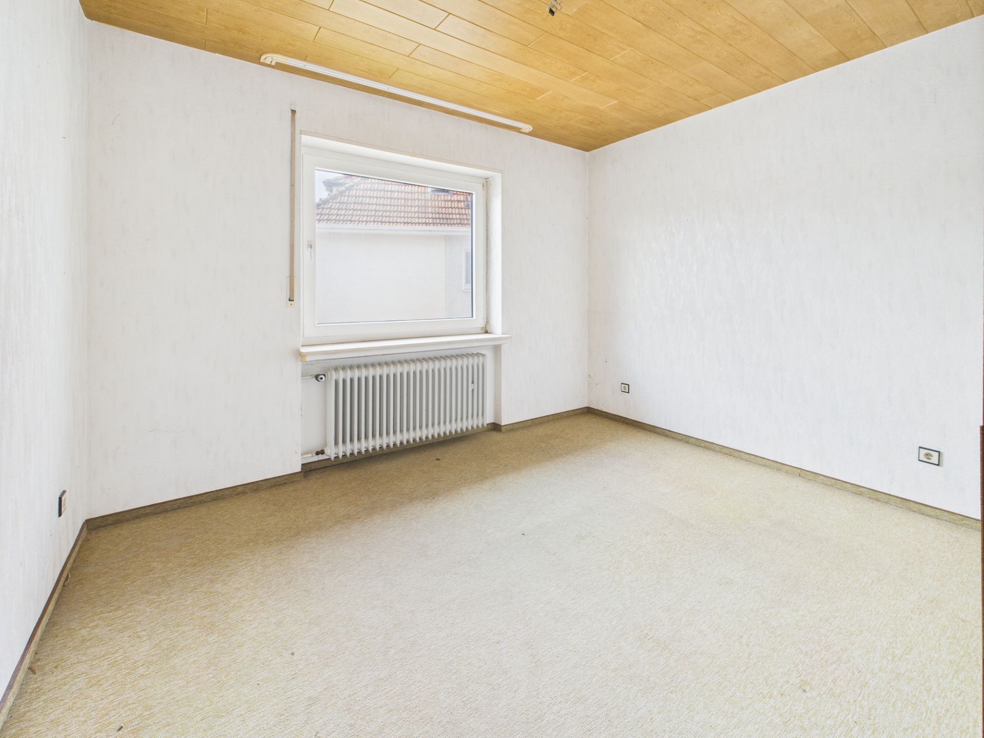Leerer Raum mit hellem Fenster, Heizkörper, weißer Wand und Holzdecke.