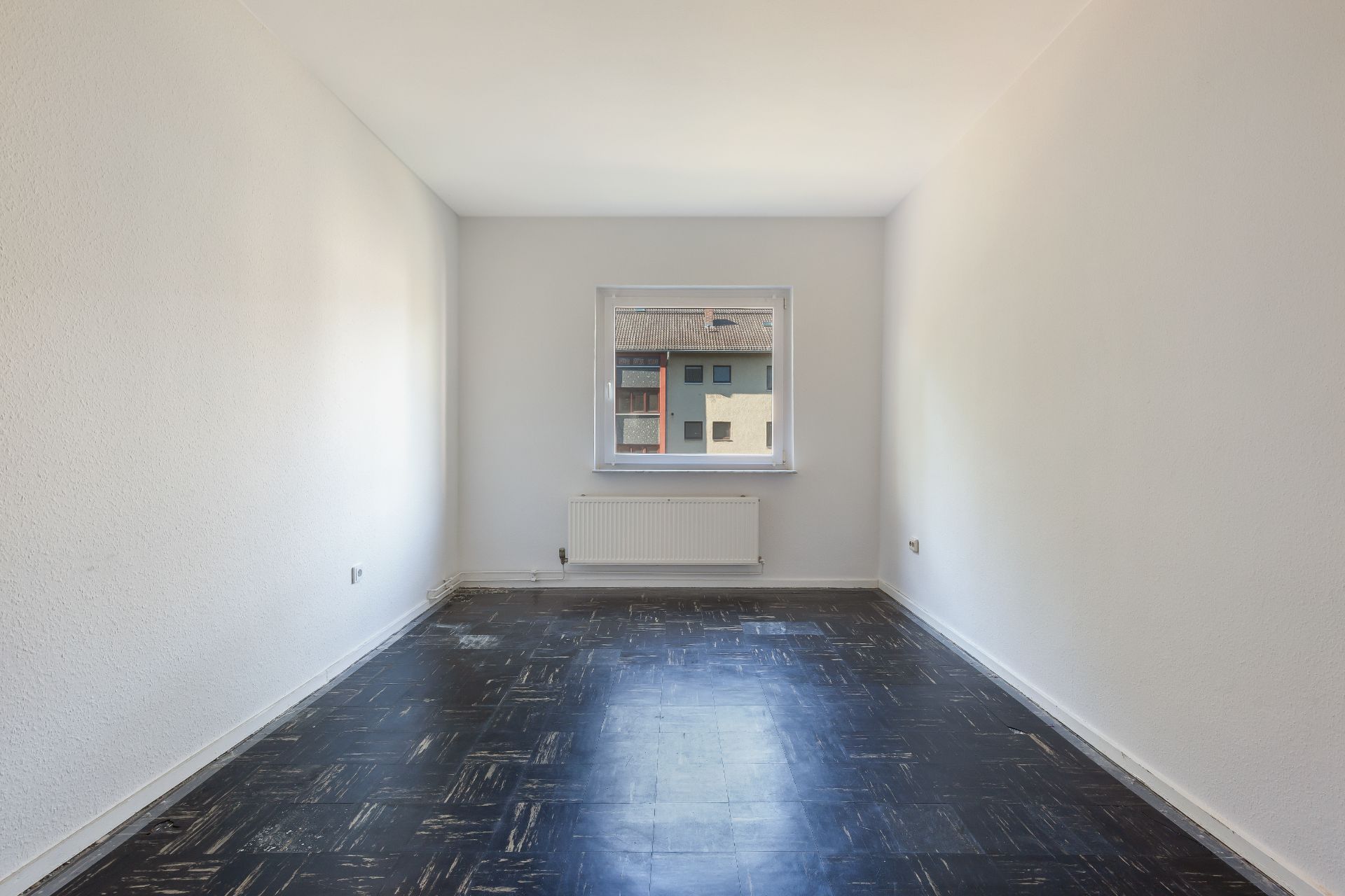 Leerer Raum mit dunklem Boden, weiß gestrichenen Wänden und Fenster mit Blick nach draußen.