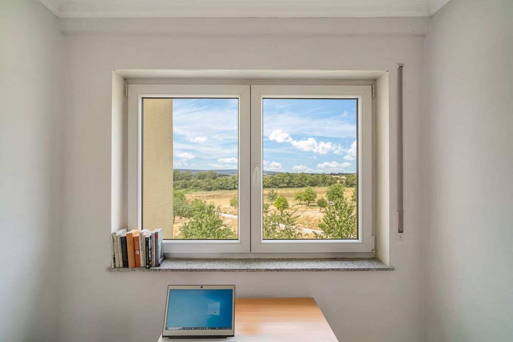 Fenster mit Blick auf Felder, Bücher auf der Fensterbank, Laptop auf Schreibtisch.