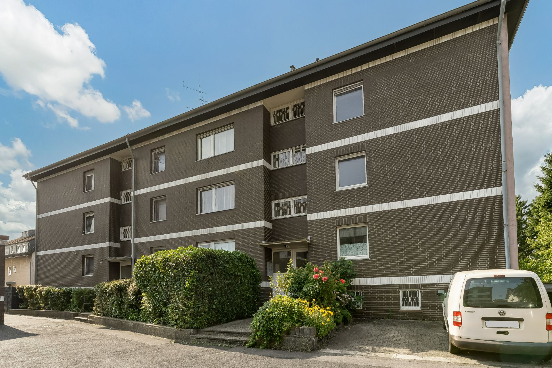 Dreistöckiges Wohnhaus mit dunkler Fassade, Büschen und geparktem weißen Auto vorne.