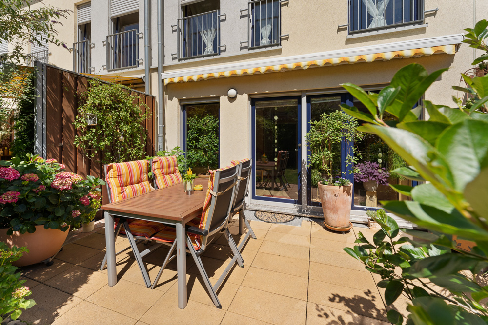 Sonnige Terrasse mit Tisch, Stühlen, Pflanzen und Blick auf eine Hausfassade mit großen Fenstern.