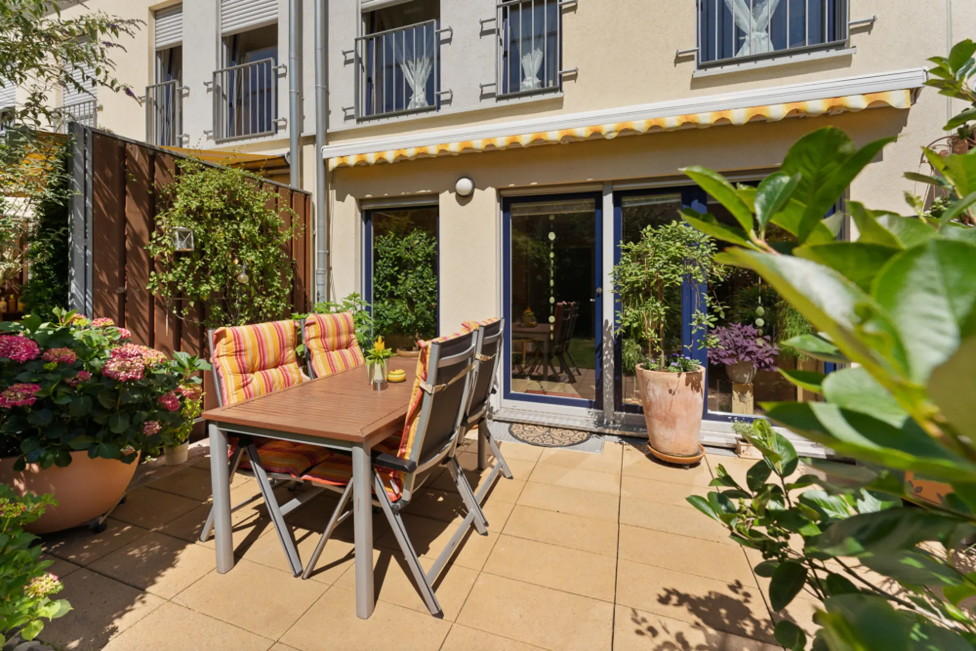 Sonnige Terrasse mit Tisch, Stühlen, Pflanzen und Blick auf eine Hausfassade mit großen Fenstern.