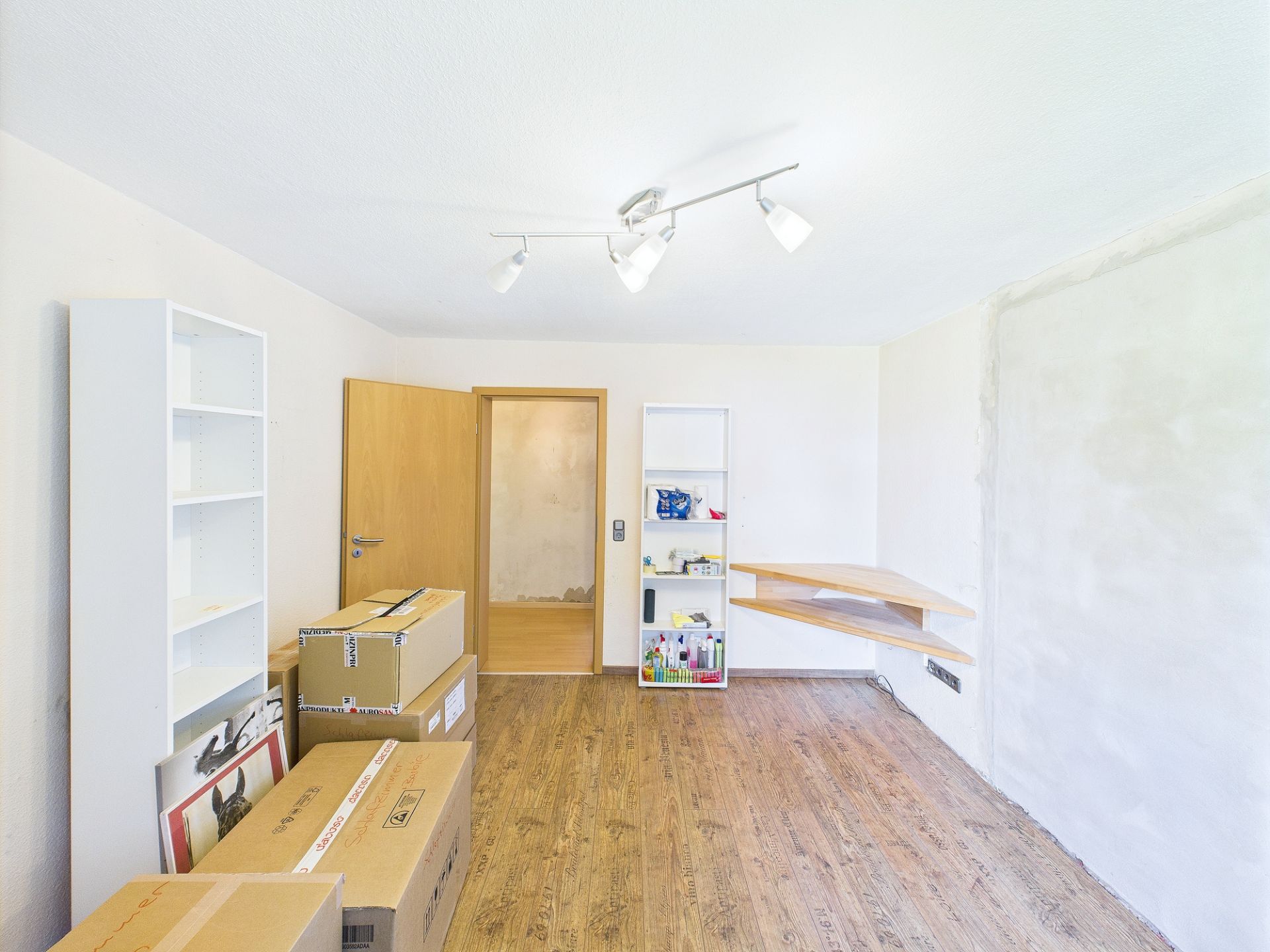 Helles Zimmer mit Holzboden, Regalen, Kartons und offenem Türrahmen.
