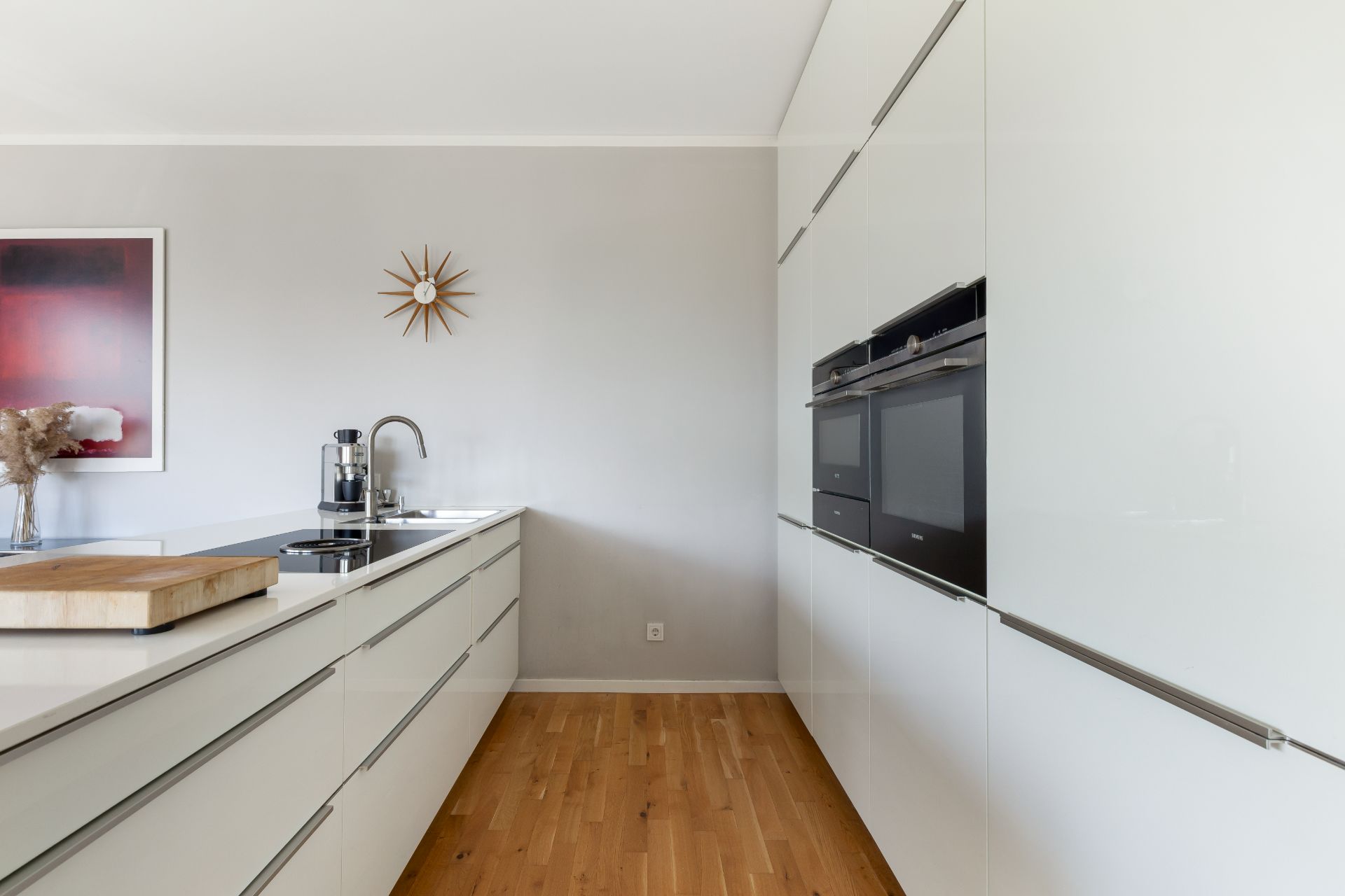 Moderne weiße Küche mit Holzfußboden, Kochinsel, Wandöfen und Deko an der Wand.