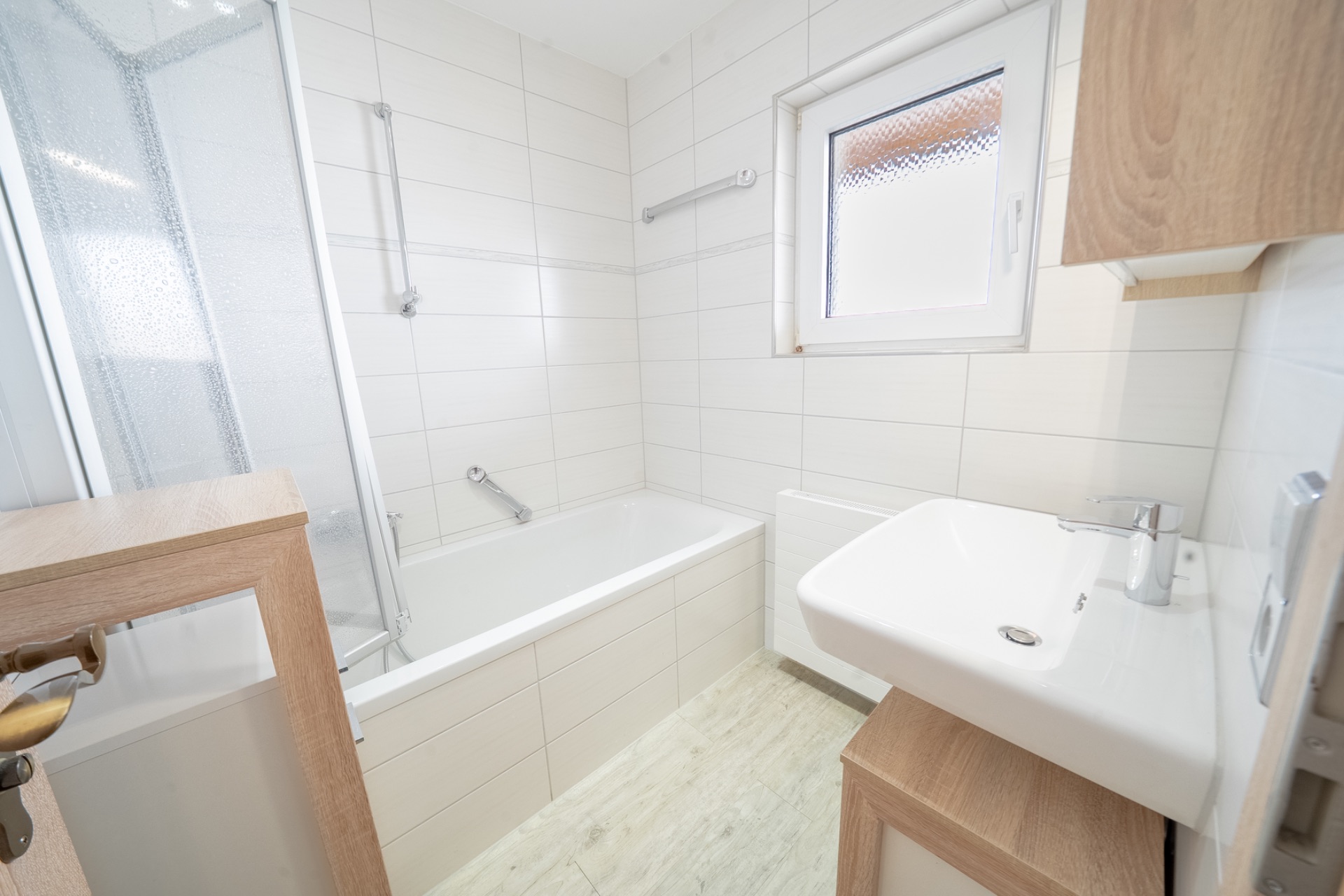 Helles Badezimmer mit Badewanne, Waschbecken, Fenster und hellen Fliesen.