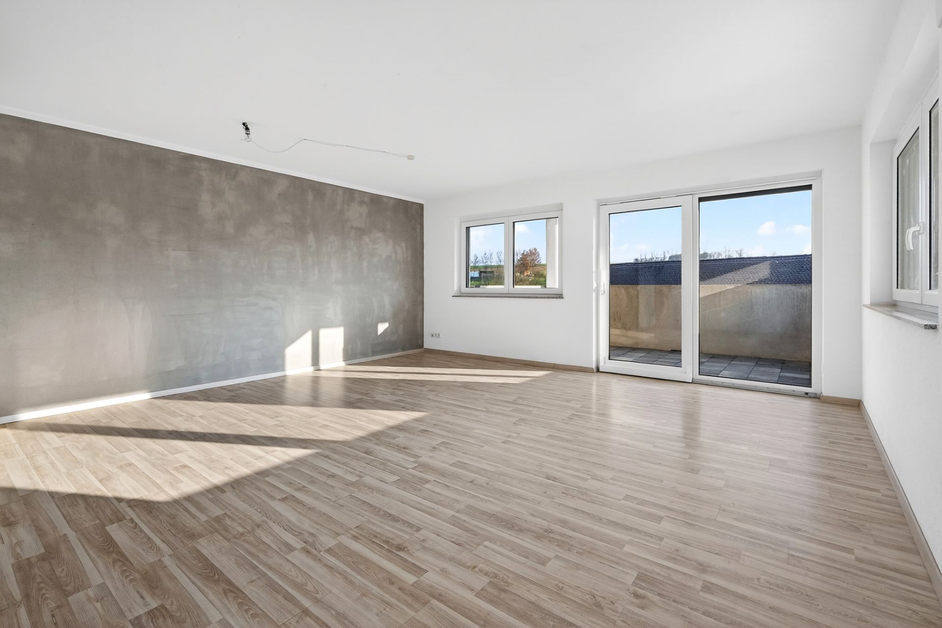 Helles, leeres Zimmer mit Parkettboden, großen Fenstern und Zugang zu einem Balkon.