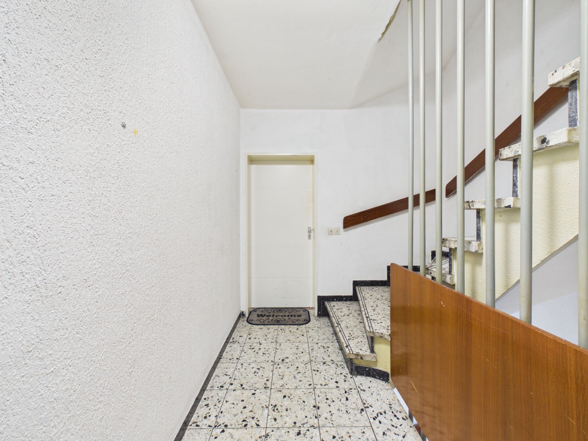 Heller Flur mit gemustertem Boden, weißer Tür und Treppe mit Holzgeländer.