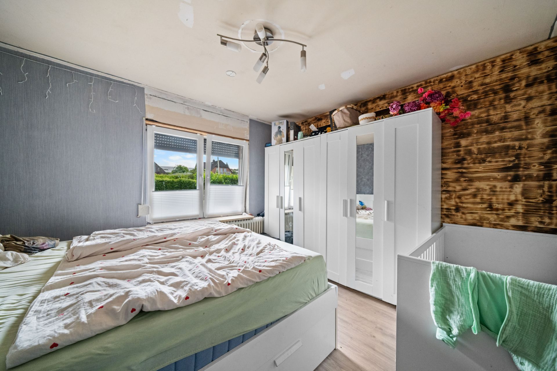 Schlafzimmer mit ungemachtem Bett, weißem Kleiderschrank, Gitterbett und Fenster mit Blick nach draußen.