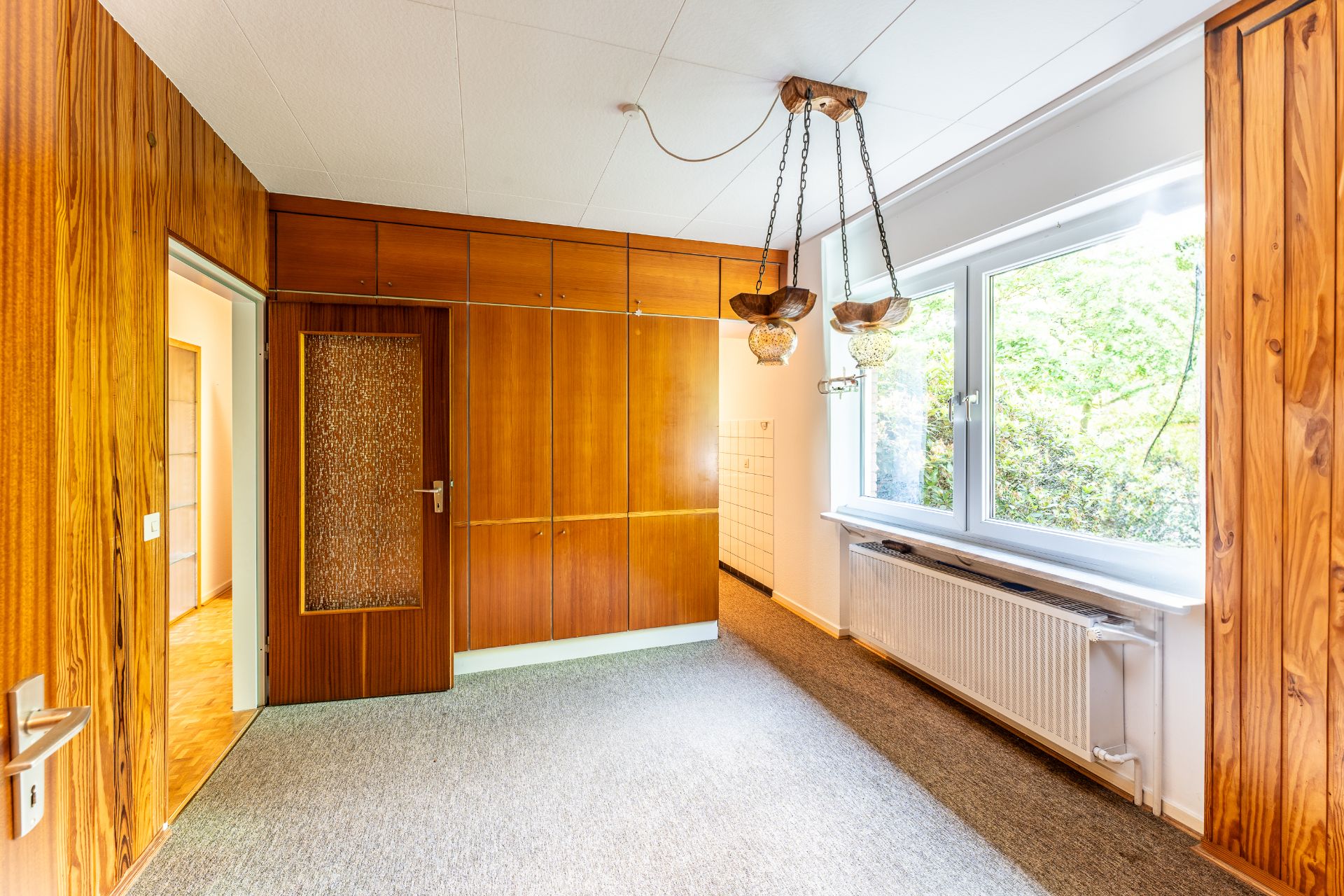 Zimmer mit Holzvertäfelung, großem Fenster, Hängelampen und Einbauschrank.