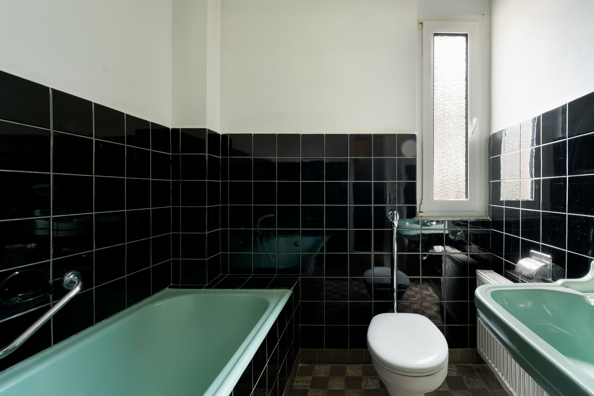 Badezimmer mit türkisfarbener Badewanne, Waschbecken, schwarzer Fliesenwand und kleinem Fenster.