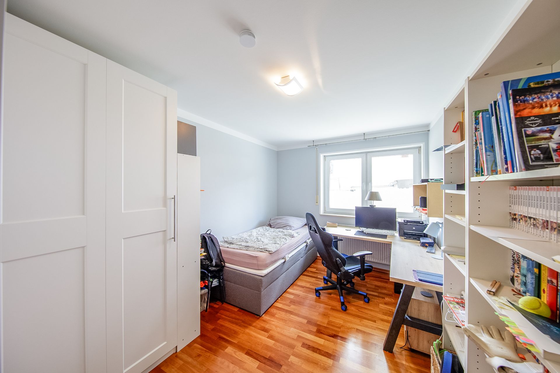 Helles Schlafzimmer mit Schreibtisch, Bett, Kleiderschrank und Regal mit Büchern.