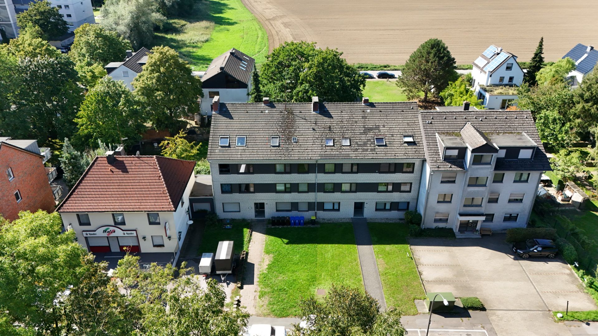 Luftaufnahme eines Mehrfamilienhauses mit Vorgarten und Parkplätzen, umgeben von Bäumen und Nachbarhäusern.